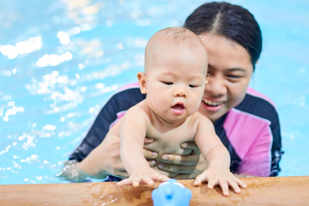 Cute little 6 months old Asian baby boy child try to grabbing a toy at the edge of indoor swimming pool, Mother and baby boy son playing, Infant Swim Classes, Baby's Developmental Milestones concept